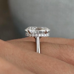 May include: A diamond ring featuring a large oval-cut central stone, surrounded by a halo of smaller diamonds. The ring has a slender, silver band. The ring is on a person's finger.