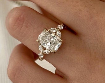 Könnte beinhalten: Ein goldener Verlobungsring mit einem großen ovalen Diamanten, der von kleineren Diamanten umgeben ist. Der Ring befindet sich an einem Finger.