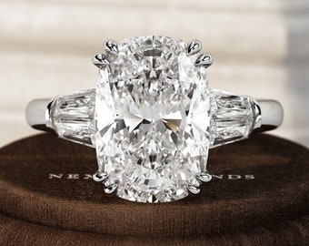 Könnte beinhalten: Ein Verlobungsring aus Platin mit einem großen, oval geschliffenen Diamanten als Mittelstein, flankiert von zwei kleineren, rechteckigen Diamanten. Der Ring liegt auf einer braunen Samtoberfläche, die das Funkeln der Edelsteine hervorhebt. Das Design des Rings ist klassisch und elegant.