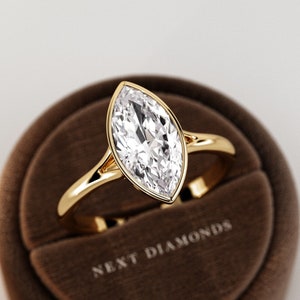 May include: A marquise-cut diamond ring with a gold band, set in a brown velvet ring box. The ring features a large, clear diamond with a pointed oval shape. The ring box has the text "NEXT DIAMONDS".