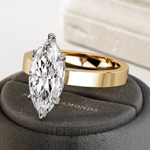 以下が含まれることがあります： 大きなクリアなマルキーズ型のダイヤモンドがついたゴールドの婚約指輪。指輪は、「Diamonds」と書かれたグレーのベルベット製の指輪箱の上に置かれています。