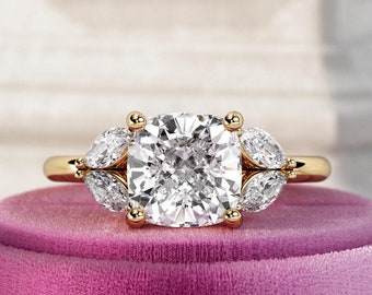 Könnte beinhalten: Ein goldener Verlobungsring mit einem großen, quadratischen Diamanten als Mittelstück und drei kleineren birnenförmigen Diamanten auf jeder Seite. Der Ring liegt auf einer rosa Samtbox.