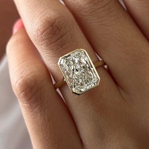 Op de afbeelding: Een rechthoekige diamanten ring met een gouden band. De diamant heeft een heldere, sprankelende uitstraling. De ring is gezet in een gouden bezel-zetting, wat een moderne en elegante uitstraling creëert. De ring zit om een vinger.