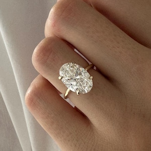 Könnte beinhalten: Ein goldener Ring mit einem großen, oval geschliffenen Diamanten. Der Diamant wird von vier Krappen gehalten, und das Band ist aus poliertem Gold. Der Ring wird an einem Finger gezeigt.