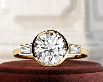 May include: A gold ring with a large, round, clear gemstone in the center, surrounded by a gold bezel. Two smaller, rectangular gemstones are set on either side of the main stone. The ring is displayed on a dark red velvet surface.