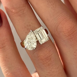 May include: A gold ring with two diamonds. The larger diamond is pear-shaped and the smaller diamond is emerald-cut.