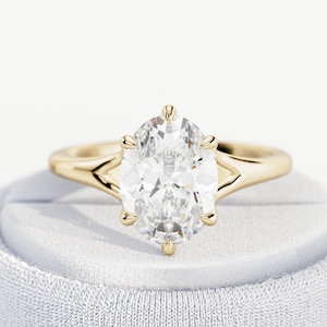 May include: A gold ring featuring a large, clear, oval-cut center stone held in place by six prongs. The band has a smooth, polished finish and a split-shank design that tapers toward the setting, creating a clean and refined appearance.