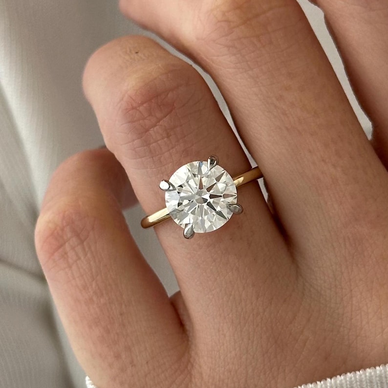 Pu&ograve; includere: Primo piano di un anello di fidanzamento in oro con un grande diamante rotondo e trasparente.