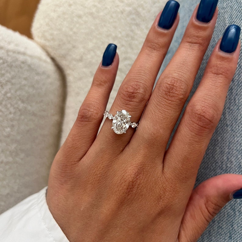 K&ouml;nnte beinhalten: Ein silberner Verlobungsring mit einem oval geschliffenen Diamanten und kleineren Akzentsteinen. Der Ring befindet sich an einer Hand mit dunkelblauem Nagellack, vor einem wei&szlig;en und blauen Hintergrund.