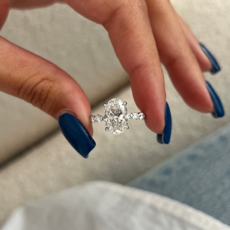 K&ouml;nnte beinhalten: Ein silberner Verlobungsring mit einem oval geschliffenen Diamanten als Mittelstein. Das Band ist mit kleineren, runden Diamanten verziert. Der Ring wird vor einem weichen Hintergrund gehalten, um sein Funkeln und Design hervorzuheben.