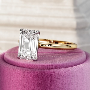 May include: A gold engagement ring with a large, clear emerald-cut diamond set in a white metal setting. The ring is sitting in a pink velvet ring box.