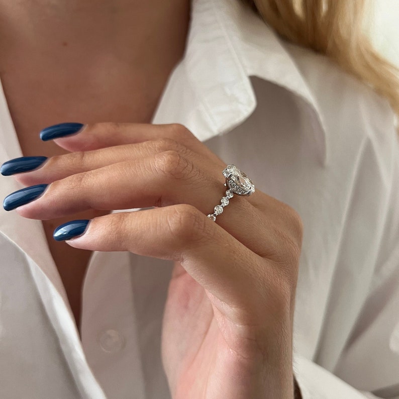 K&ouml;nnte beinhalten: Ein silberner Ring mit einem gro&szlig;en, oval geschliffenen Diamanten als Mittelstein und kleineren runden Diamanten entlang des Rings. Der Ring ist vor einem wei&szlig;en Hintergrund abgebildet.