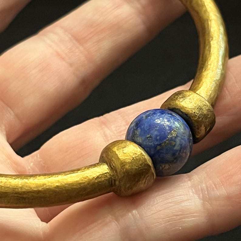 May include: A gold-coloured bracelet with a textured finish. The bracelet features a round, deep blue bead at the centre, flanked by two gold-coloured, textured accents. The bracelet is a piece of jewellery.