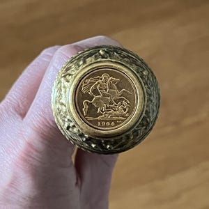 May include: A gold coin featuring a man on horseback, dated 1904, set within a textured, round, gold-toned object. The object is held in a hand, with a blurred wooden background.