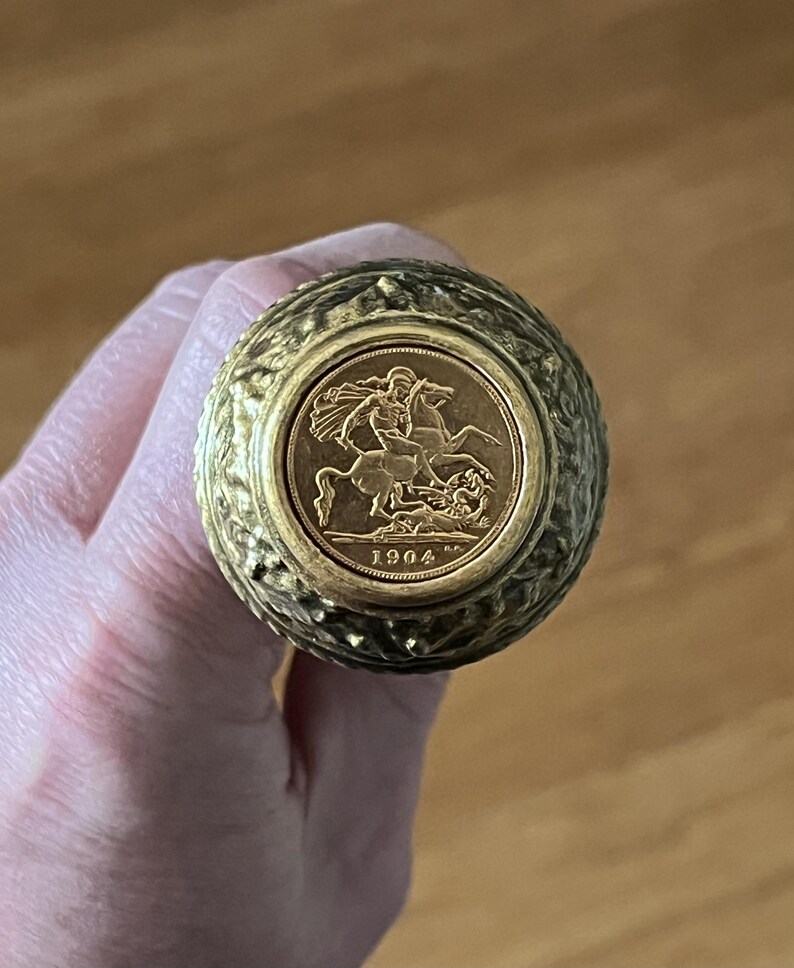 May include: A gold coin featuring a man on horseback, dated 1904, set within a textured, round, gold-toned object. The object is held in a hand, with a blurred wooden background.