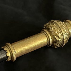 May include: A detailed view of a golden ceremonial scepter. The scepter has ornate embossed designs and a cylindrical shaft. The top and base are decorated with intricate patterns, possibly indicating a historical or royal context. The background is dark.