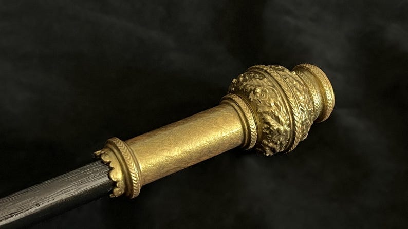 May include: A detailed view of a golden ceremonial scepter. The scepter has ornate embossed designs and a cylindrical shaft. The top and base are decorated with intricate patterns, possibly indicating a historical or royal context. The background is dark.