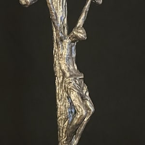 May include: A silver-toned crucifix sculpture depicting a figure on a cross. The sculpture has a textured, weathered appearance, with the figure's form detailed. The cross is made to look like a tree trunk.