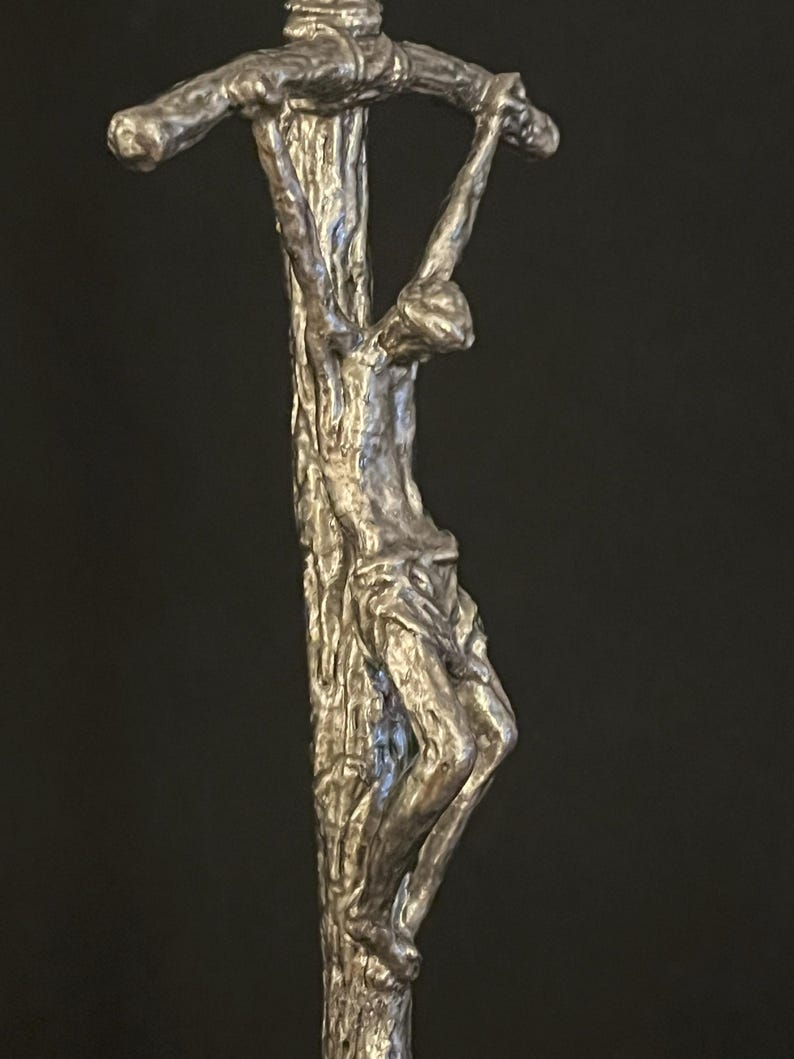 May include: A silver-toned crucifix sculpture depicting a figure on a cross. The sculpture has a textured, weathered appearance, with the figure's form detailed. The cross is made to look like a tree trunk.