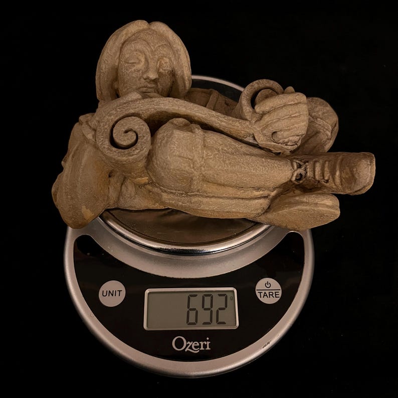 May include: A beige sculpture of a person lying down, with closed eyes and a serene expression. The figure is resting on a digital scale, which displays a weight of 6.92. The scale is silver and black, with the brand name "Ozeri" visible.