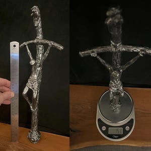 May include: A silver-colored crucifix sculpture with a detailed depiction of Jesus on the cross. The sculpture is approximately 12 inches tall and has a textured, weathered appearance. The cross is held by a person's hand.