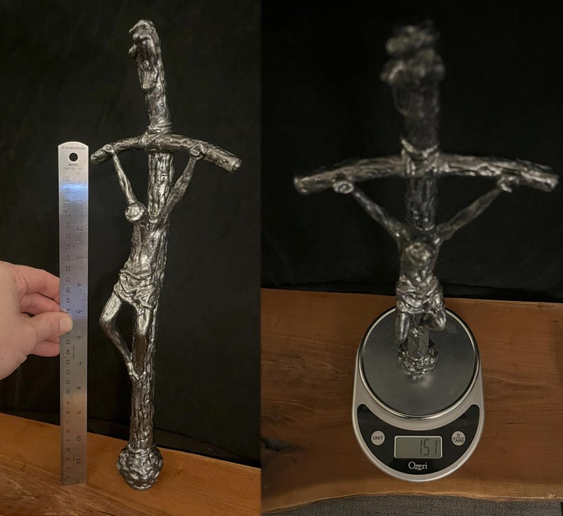May include: A silver-colored crucifix sculpture with a detailed depiction of Jesus on the cross. The sculpture is approximately 12 inches tall and has a textured, weathered appearance. The cross is held by a person's hand.