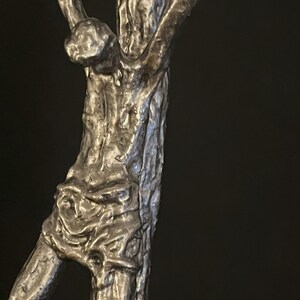 May include: A detailed, close-up view of a silver-toned sculpture depicting a figure on a cross. The sculpture has a textured, rough surface, with visible details of the body and the cross. The background is a solid black.