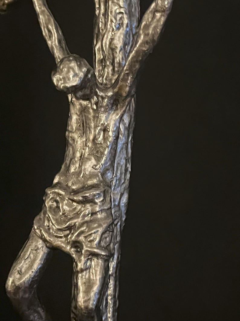 May include: A detailed, close-up view of a silver-toned sculpture depicting a figure on a cross. The sculpture has a textured, rough surface, with visible details of the body and the cross. The background is a solid black.