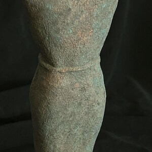 May include: A green metal sculpture of a person with their arms raised above their head. The sculpture has a textured surface and a belt around the waist.