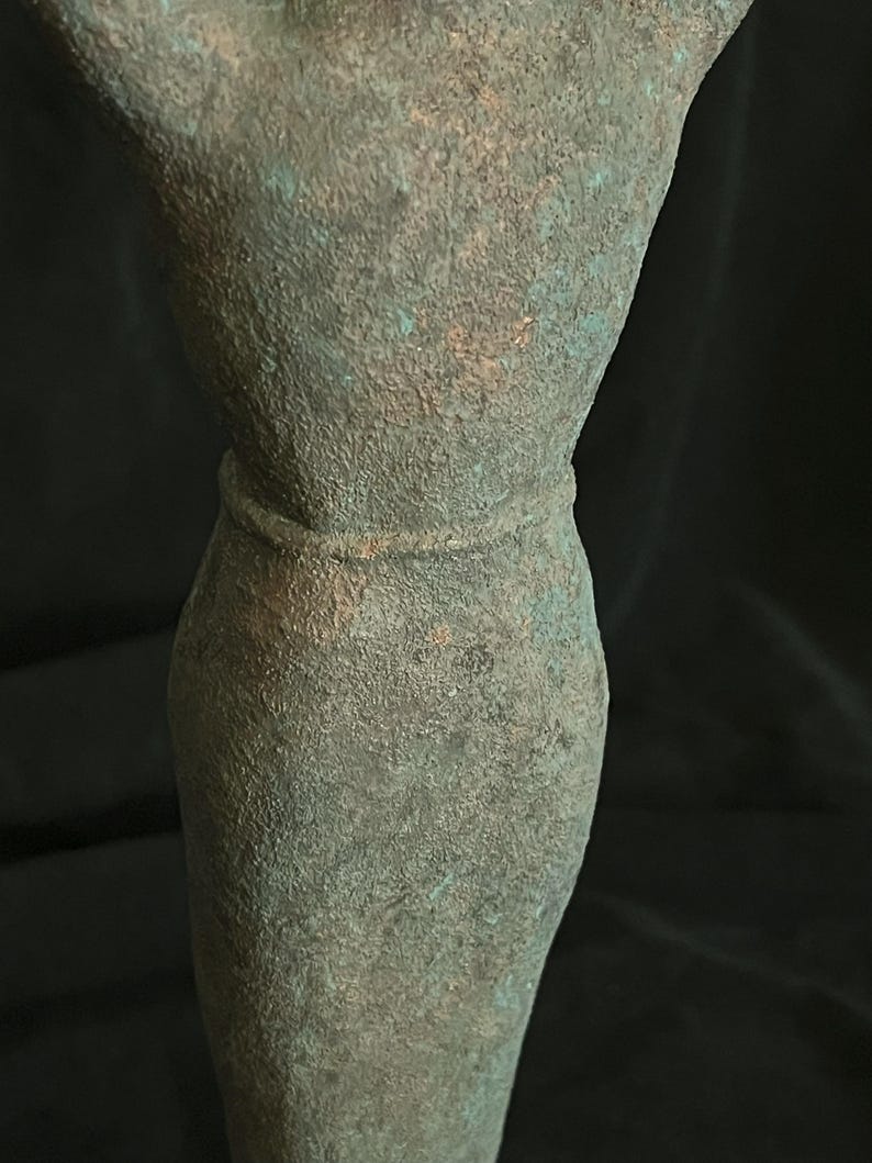 May include: A green metal sculpture of a person with their arms raised above their head. The sculpture has a textured surface and a belt around the waist.