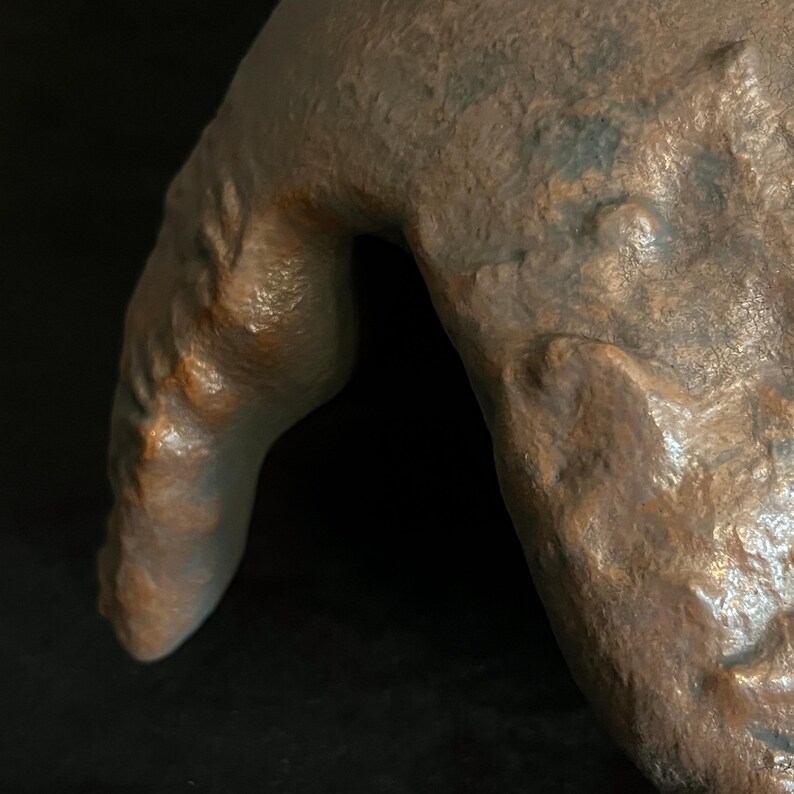 Puede incluir: Primer plano de una escultura de color bronce de un brazo y una mano humanos. La escultura tiene una superficie rugosa y texturizada con detalles visibles de venas y piel. La iluminaci&oacute;n crea sombras que enfatizan la forma y la textura.