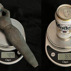 May include: Two digital kitchen scales with a bronze-colored statue of a woman on one scale and a can of Modelo Especial beer on the other scale. The scales display the weight of each item in ounces.