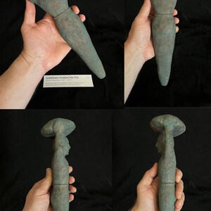 May include: A green stone sculpture of a figure holding a round object above their head. The sculpture is about 15 inches tall and is made of a smooth, green stone. The figure is standing on a pedestal and is wearing a long, flowing robe. The sculpture is a Sumerian foundation peg, also known as a "peg-figurine".