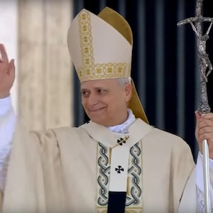 May include: A person wearing a cream-colored religious garment, waving with a gold ring on their finger. They hold a silver staff with a crucifix. The garment has decorative trim and a tall, pointed hat.