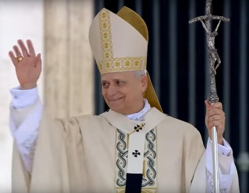 May include: A person wearing a cream-colored religious garment, waving with a gold ring on their finger. They hold a silver staff with a crucifix. The garment has decorative trim and a tall, pointed hat.