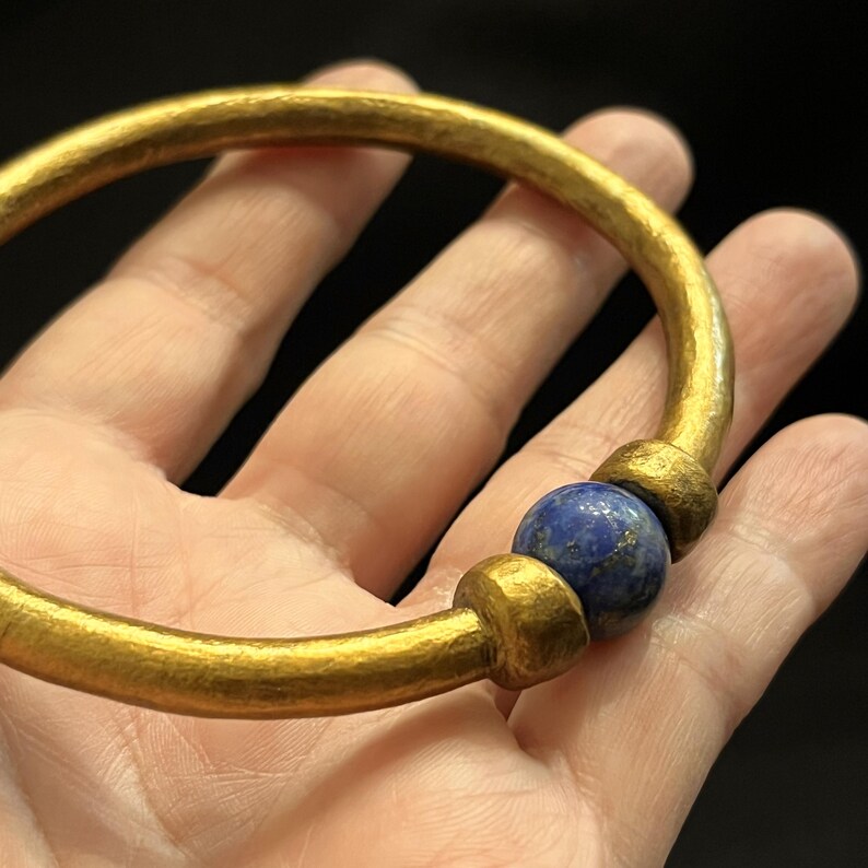 May include: A gold-coloured bangle bracelet with a hammered texture. The bracelet features a blue lapis lazuli bead and two gold-coloured accent beads. The bracelet is a simple, elegant design.