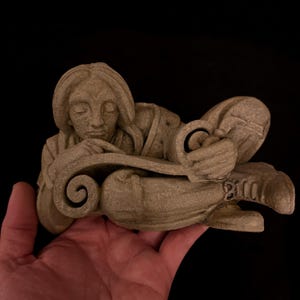 May include: A beige-colored sculpture of a reclining figure with closed eyes, resting on a scroll. The figure is holding the scroll with both hands. The sculpture is held in a person's hand against a black background.