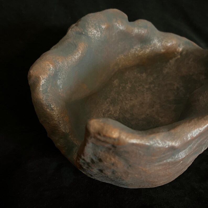 Puede incluir: Un cuenco hecho a mano, de forma org&aacute;nica, con una superficie texturizada. El cuenco presenta una mezcla de tonos bronce y gris, creando una est&eacute;tica r&uacute;stica. El interior es de un tono m&aacute;s oscuro, contrastando con los bordes m&aacute;s claros.