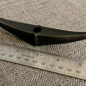 以下が含まれることがあります： 中央に穴が開いた黒い金属製のツルハシヘッド。ツルハシヘッドの長さは約10cmで、灰色の布地に置かれています。