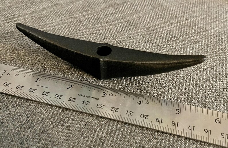 以下が含まれることがあります： 中央に穴が開いた黒い金属製のツルハシヘッド。ツルハシヘッドの長さは約10cmで、灰色の布地に置かれています。