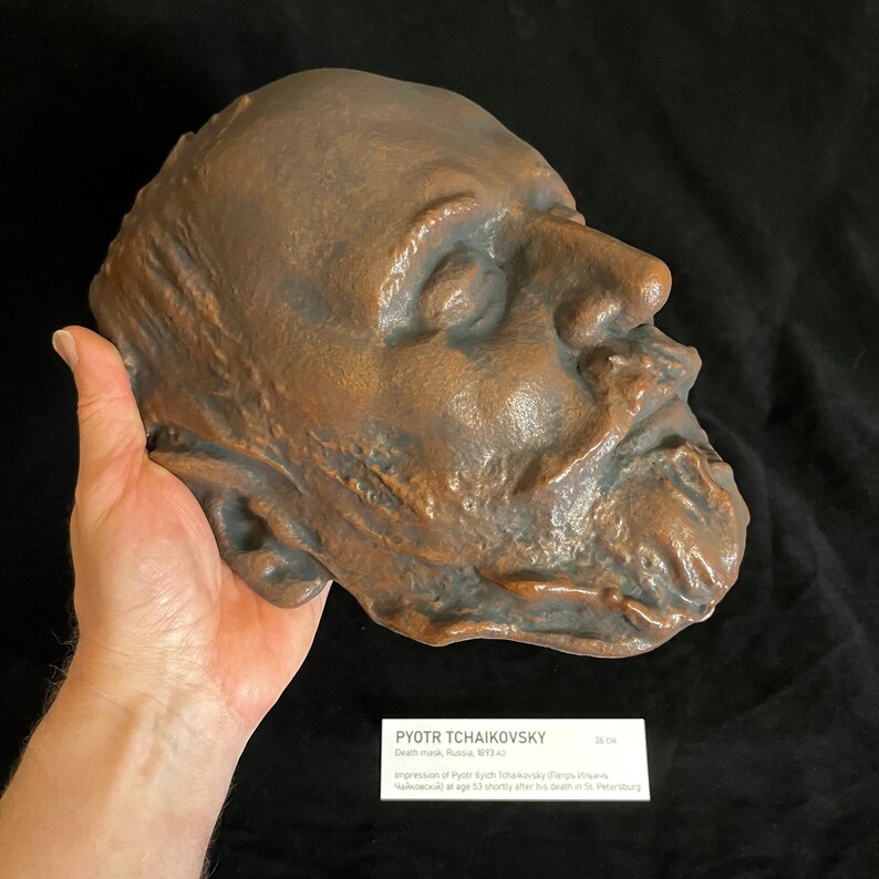 Puede incluir: Una m&aacute;scara mortuoria de color bronce de Piotr Ilich Chaikovski, con rasgos faciales detallados, incluyendo barba y arrugas. La m&aacute;scara est&aacute; sostenida en una mano. Una peque&ntilde;a tarjeta blanca con texto es visible.