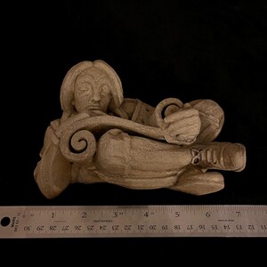 May include: A beige-colored sculpture of a reclining figure, possibly a classical depiction. The figure rests on a scroll-like form, with detailed facial features and curled elements. The sculpture is approximately 6 inches long.