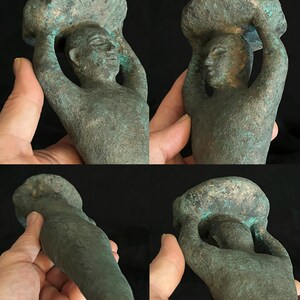May include: A green bronze sculpture of a person carrying a large object on their head. The sculpture is stylized and abstract, with the person's arms raised above their head. The sculpture is made of a smooth, green bronze.