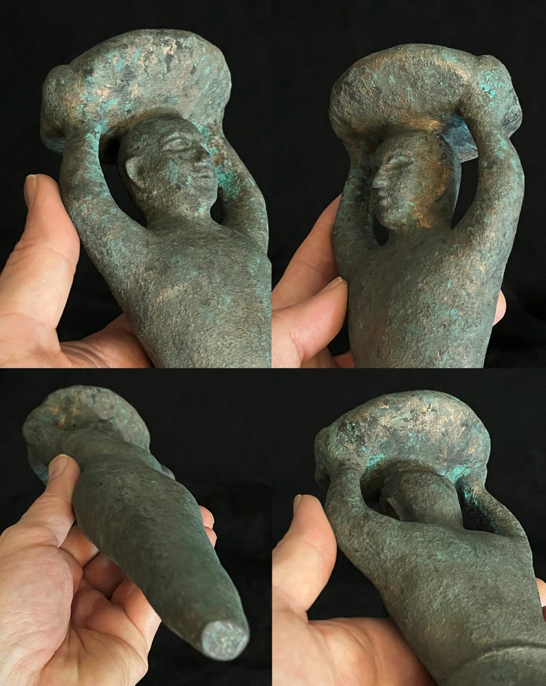May include: A green bronze sculpture of a person carrying a large object on their head. The sculpture is stylized and abstract, with the person's arms raised above their head. The sculpture is made of a smooth, green bronze.
