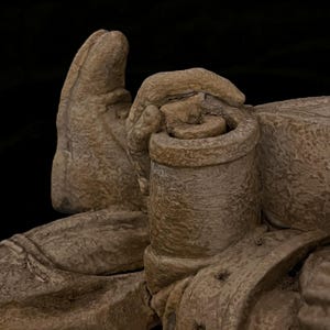 May include: A close-up of a beige-colored sculpture depicting a hand holding a cylindrical object. The sculpture has a rough, textured surface and is set against a black background. The details of the hand and the object are clearly visible.