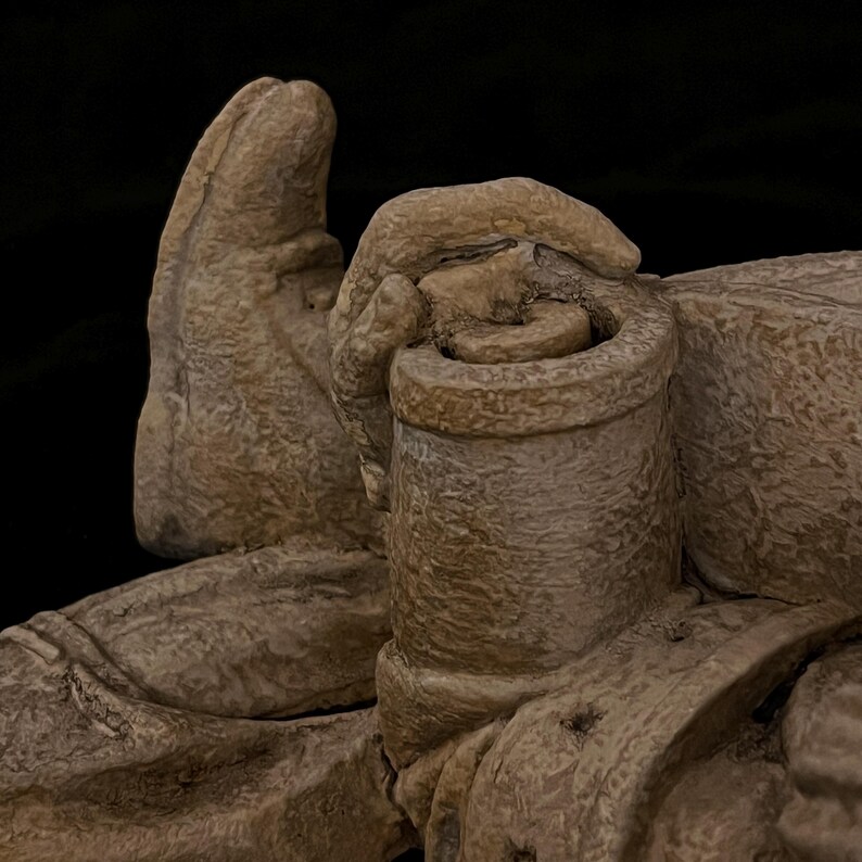 May include: A close-up of a beige-colored sculpture depicting a hand holding a cylindrical object. The sculpture has a rough, textured surface and is set against a black background. The details of the hand and the object are clearly visible.