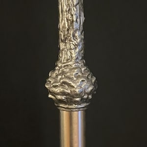 May include: Close-up of a silver-colored walking cane. The top portion features a textured, organic design, resembling tree bark. The handle has a bulbous shape, and the shaft is a smooth, polished metal.