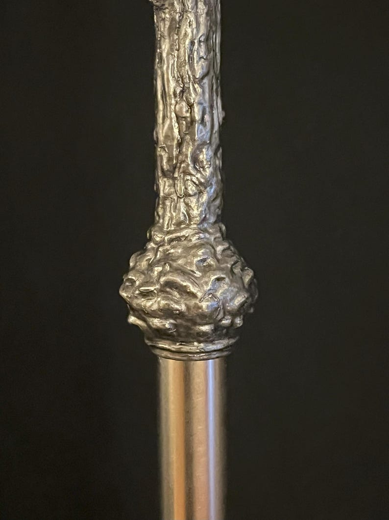 May include: Close-up of a silver-colored walking cane. The top portion features a textured, organic design, resembling tree bark. The handle has a bulbous shape, and the shaft is a smooth, polished metal.