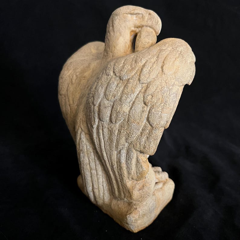 May include: A beige-colored sculpture of an eagle with detailed feather carvings. The eagle is depicted with its wings partially spread, and its head is turned to the side. The sculpture is made of a stone-like material and sits on a small base.