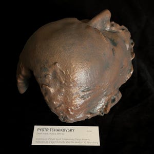 Puede incluir: Una m&aacute;scara mortuoria de color bronce de Piotr Chaikovski, creada en Rusia en 1893. La m&aacute;scara mide aproximadamente 26 cm de largo. La imagen incluye una etiqueta con el nombre del compositor y detalles sobre la creaci&oacute;n de la m&aacute;scara.